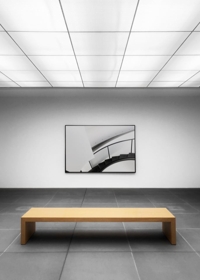Light wood stained bench positioned in front of a hanging framed artwork of an architectural image of stairs